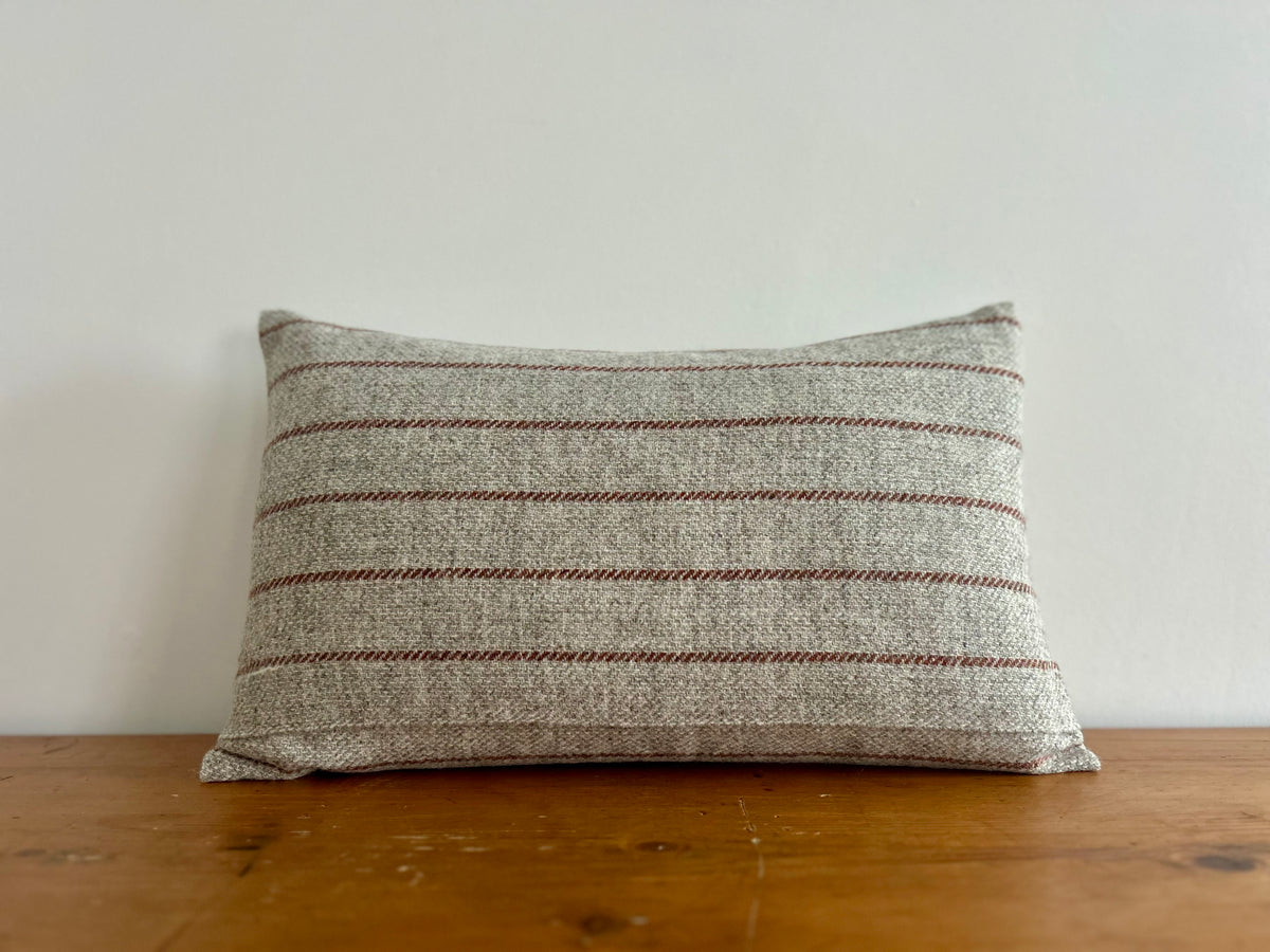 A rectangular throw pillow with a textured gray and beige woven fabric featuring thin horizontal rust-brown stripes, resting on a wooden surface against a plain white wall.